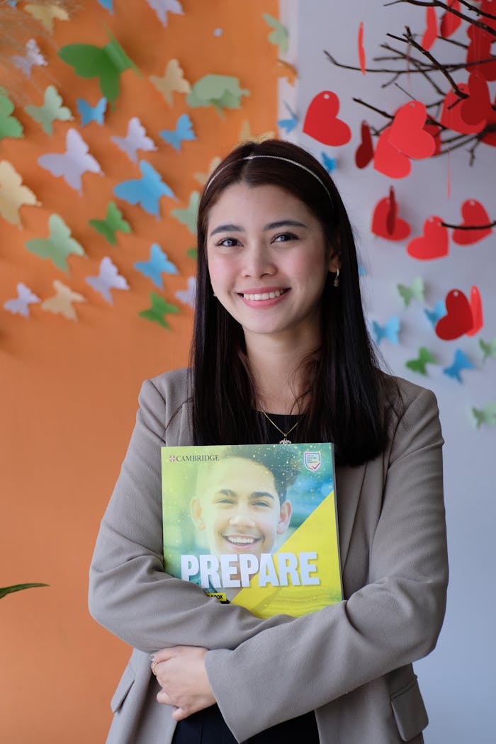 A young woman smiling while holding a Cambridge book in a classroom setting with colorful decor.